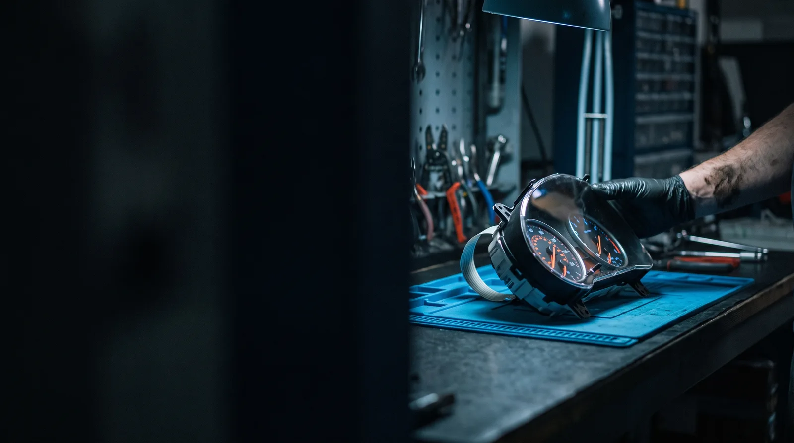 A car instrument cluster removed from the dashboard being examined on a blue ESD-safe workbench in a dark automotive electronics repair workshop