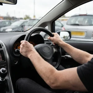 Driver's hands gripping the steering wheel of a right-hand-drive car in a UK car park, straining against heavy steering at low speed