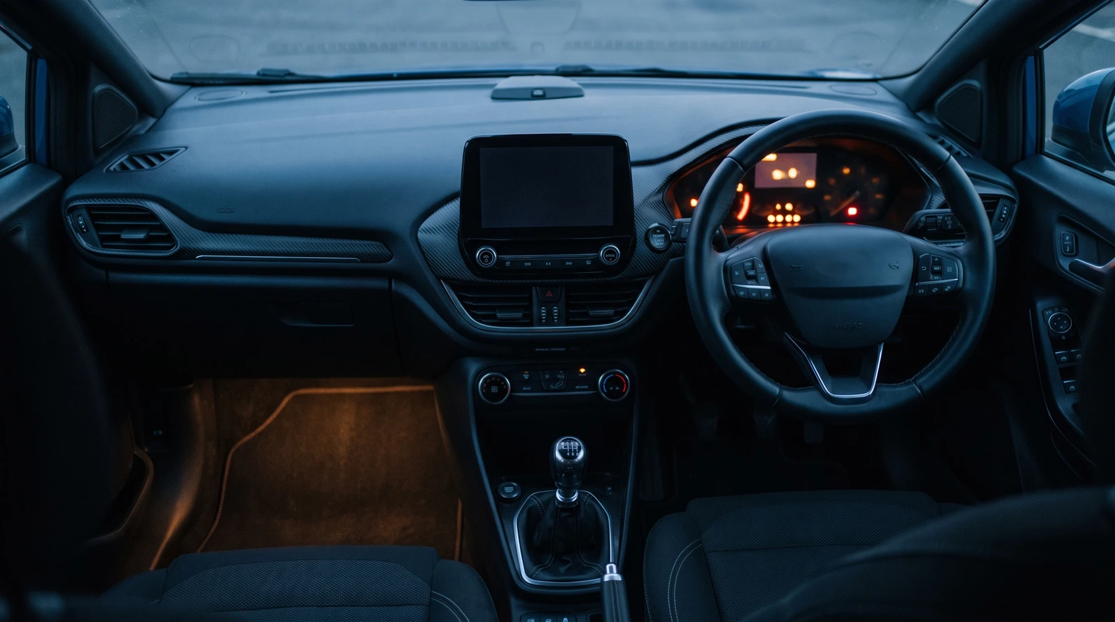 Right-hand-drive Ford Puma cabin at dusk: steering wheel on the right with the instrument cluster lit behind it, and the centre touchscreen and manual gear lever to the driver's left
