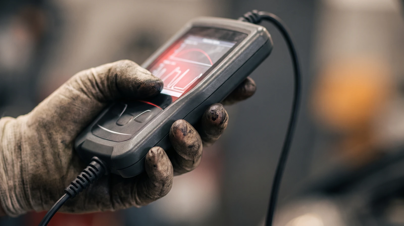 A mechanic's gloved hand holding a handheld OBD diagnostic scan tool in a workshop, its screen showing a red live-data trace, while investigating a lean-running fault